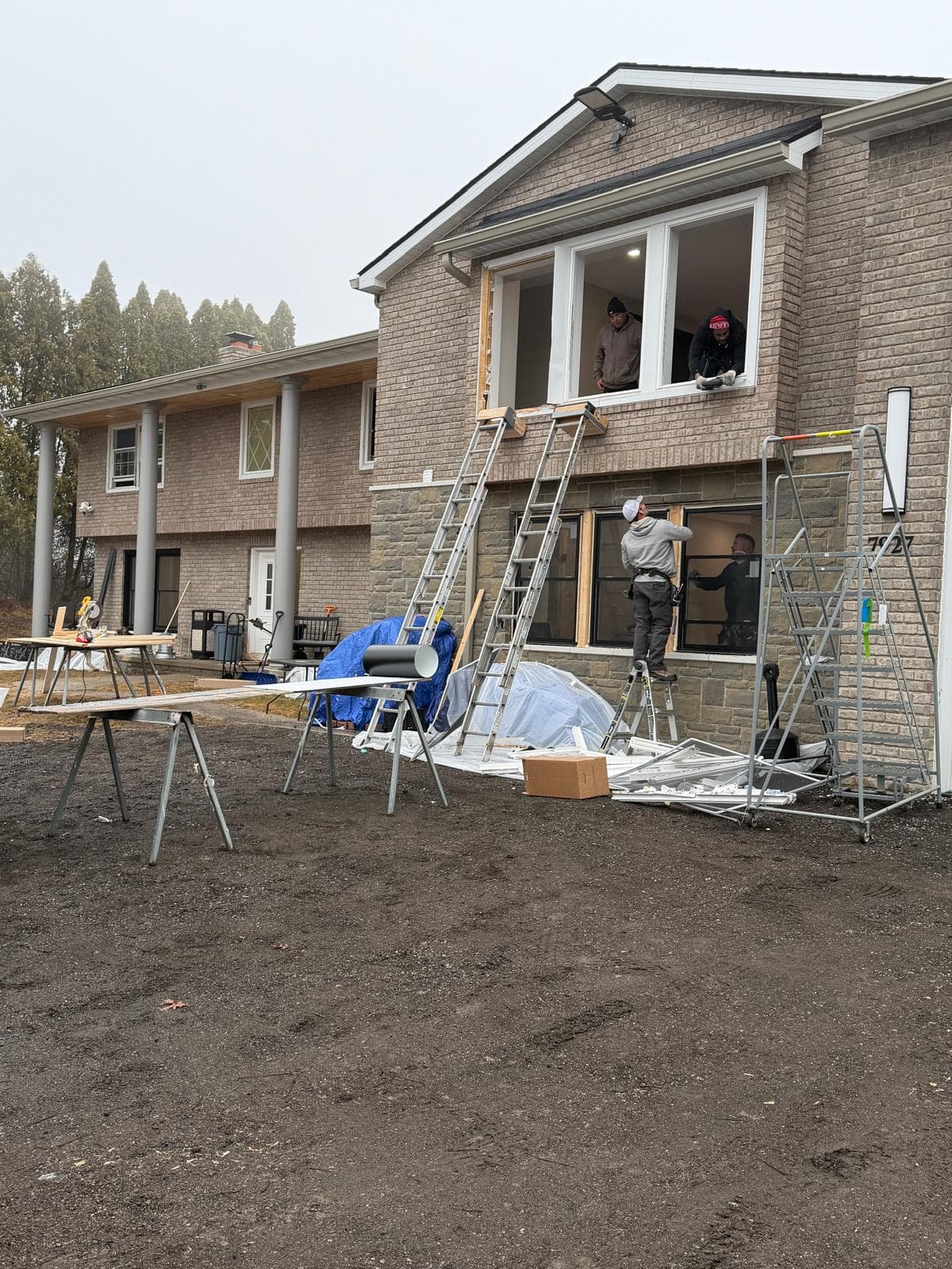 Construction workers installing new windows on a two-story brick house with ladders and equipment.