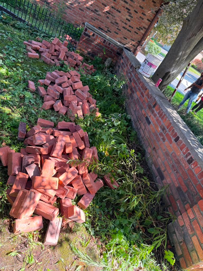 Several piles of red bricks on a grassy area next to a brick wall.