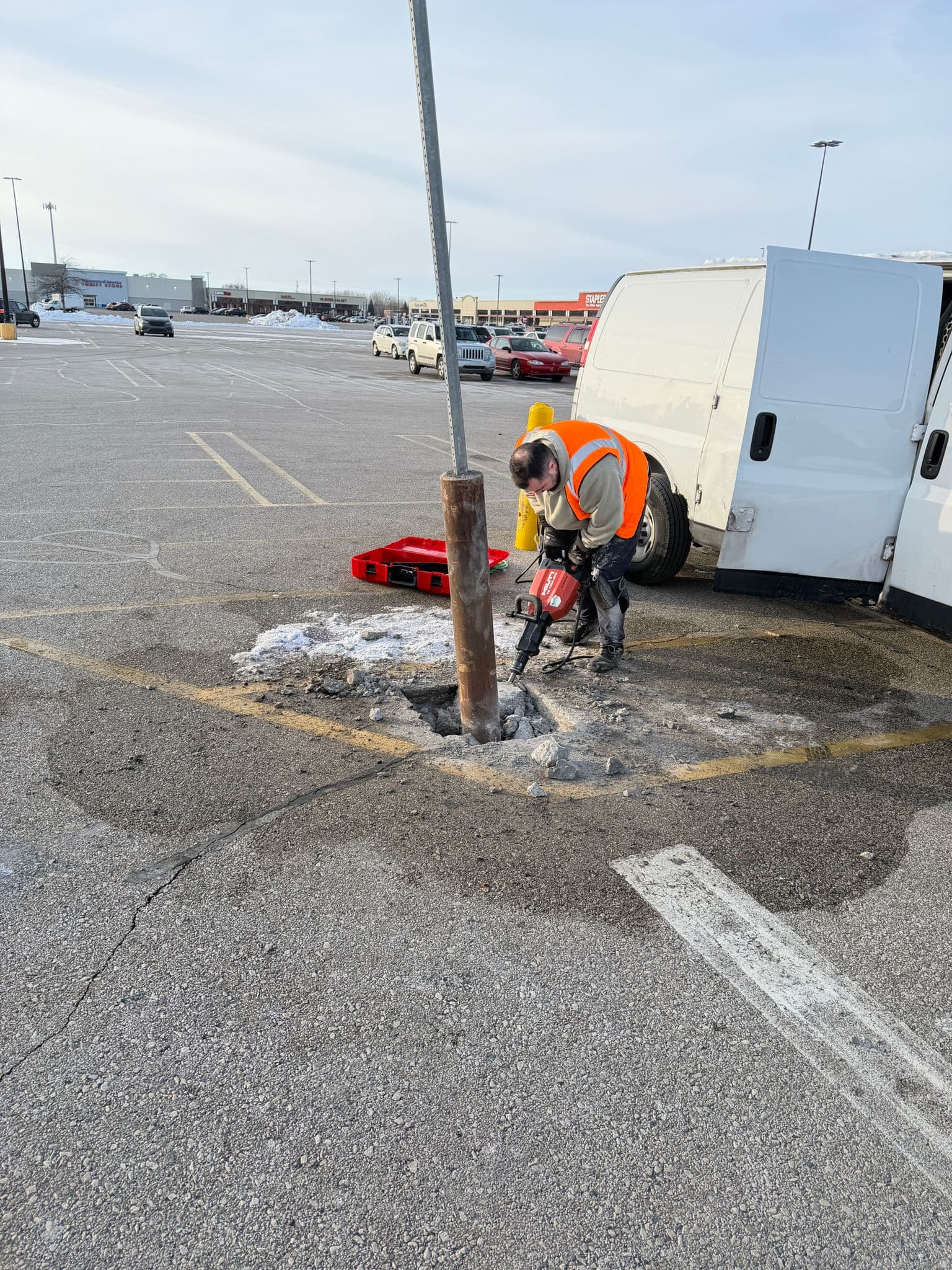 Construction worker using a jackhammer to break asphalt around a metal post in a parking lot.