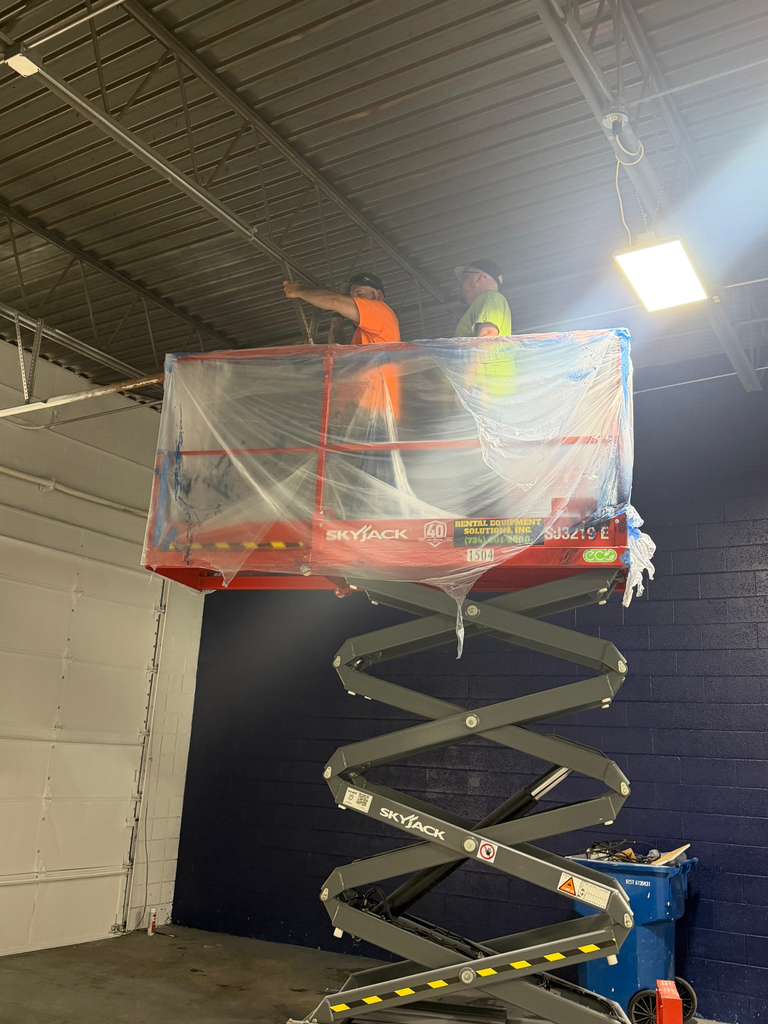 Two workers on an elevated scissor lift platform working on the ceiling of a warehouse.