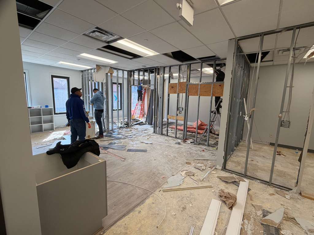 Workers renovating an office interior with exposed metal wall studs and scattered floor debris.